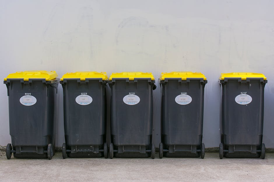 Five large wheeled rubbish bins lined up in a row against a plain, light-colored wall. The bins are made of durable black plastic with textured surfaces and fitted with yellow lids that contrast with